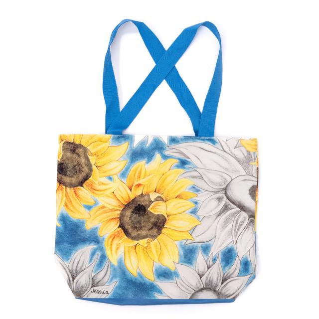 Canvas tote bag with black and white sunflowers all over, featuring one bright yellow sunflower, with blue handles.